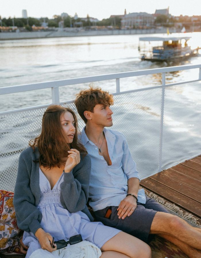 The Wade A couple sits on a wooden dock, smiling, with a boat gently floating in the background under a clear blue sky.